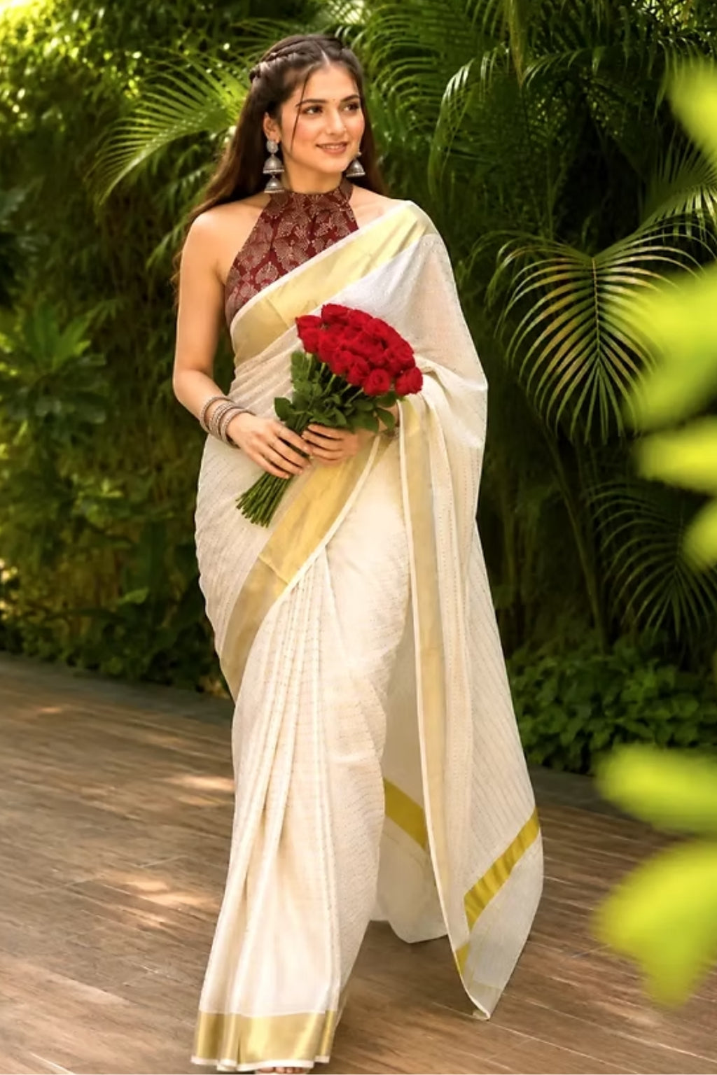 A woman wearing a white silk saree with a gold border and a dark brown patterned sleeveless blouse, holding a bouquet of red roses while standing outdoors in front of lush green foliage.