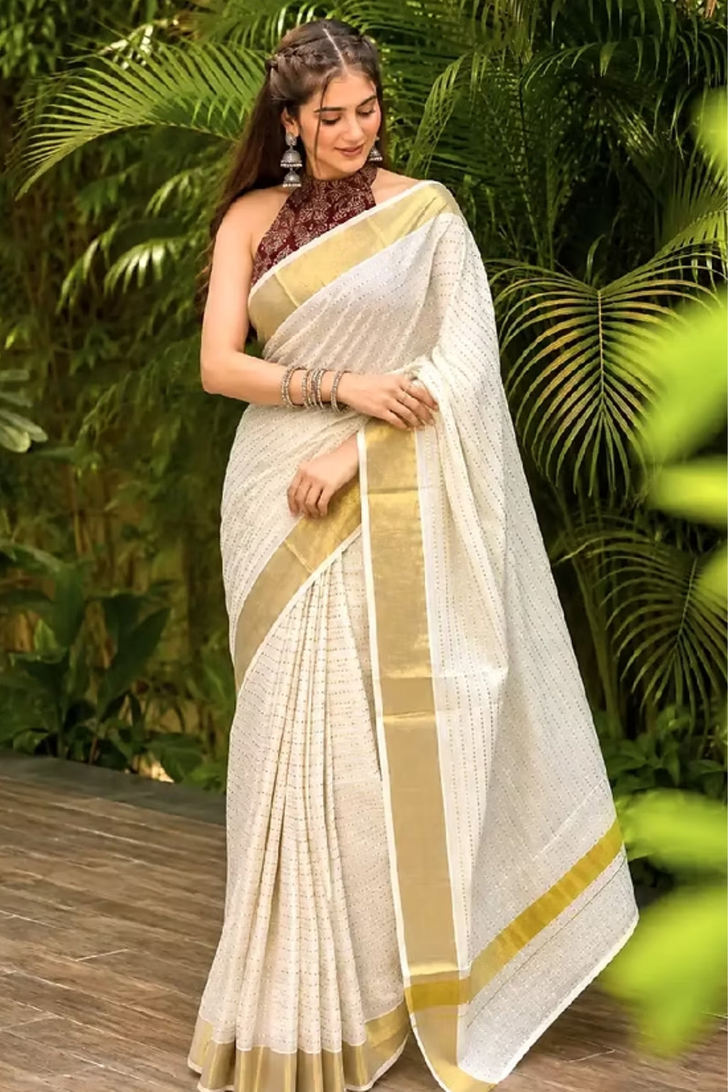 A woman wearing a cream-colored saree with a gold border and a brown patterned blouse, standing outdoors in front of lush green tropical foliage.