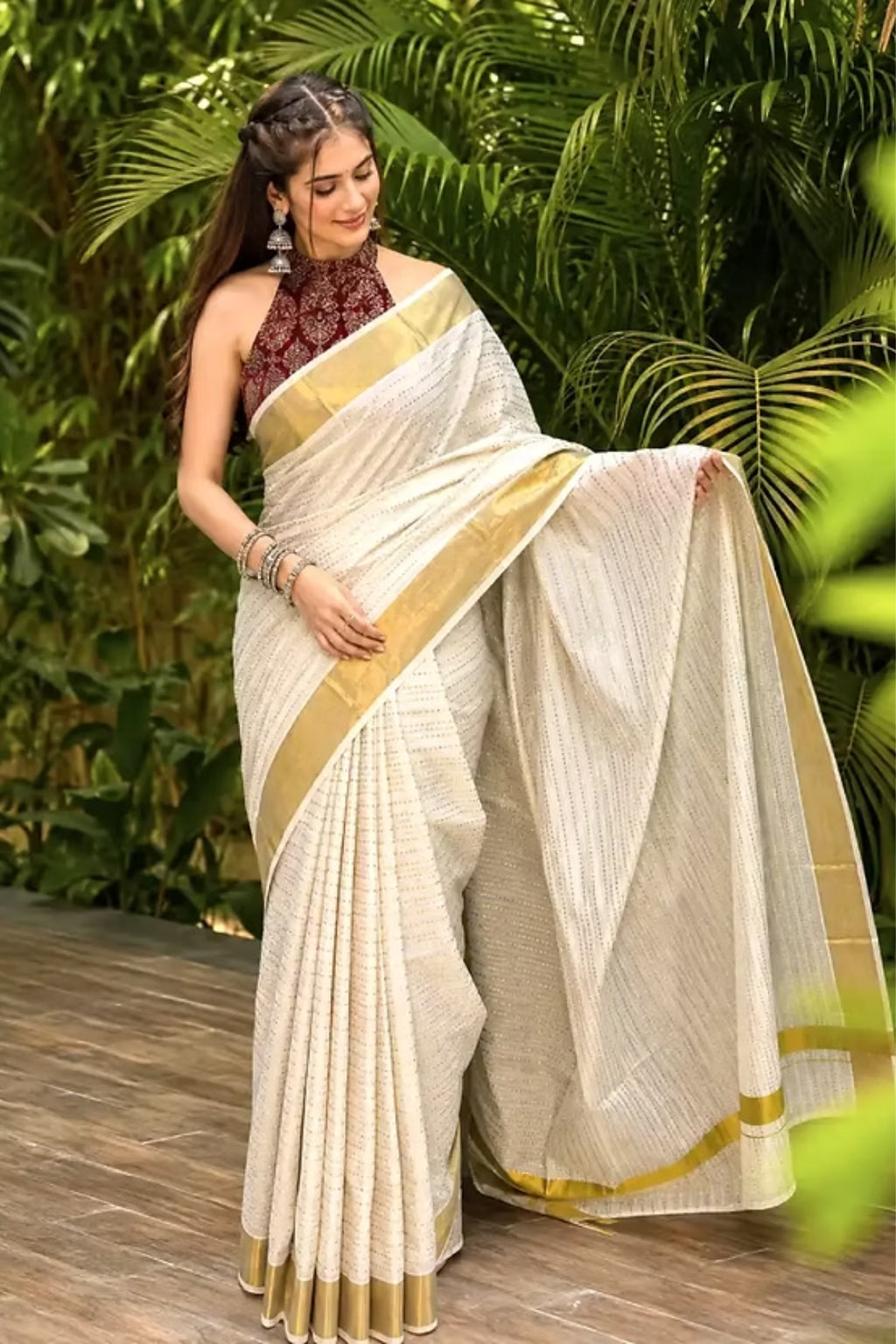 A woman models a cream-colored cotton saree with gold stripes and a matching cream dupatta, paired with a dark red patterned sleeveless blouse, set against a lush green tropical background.