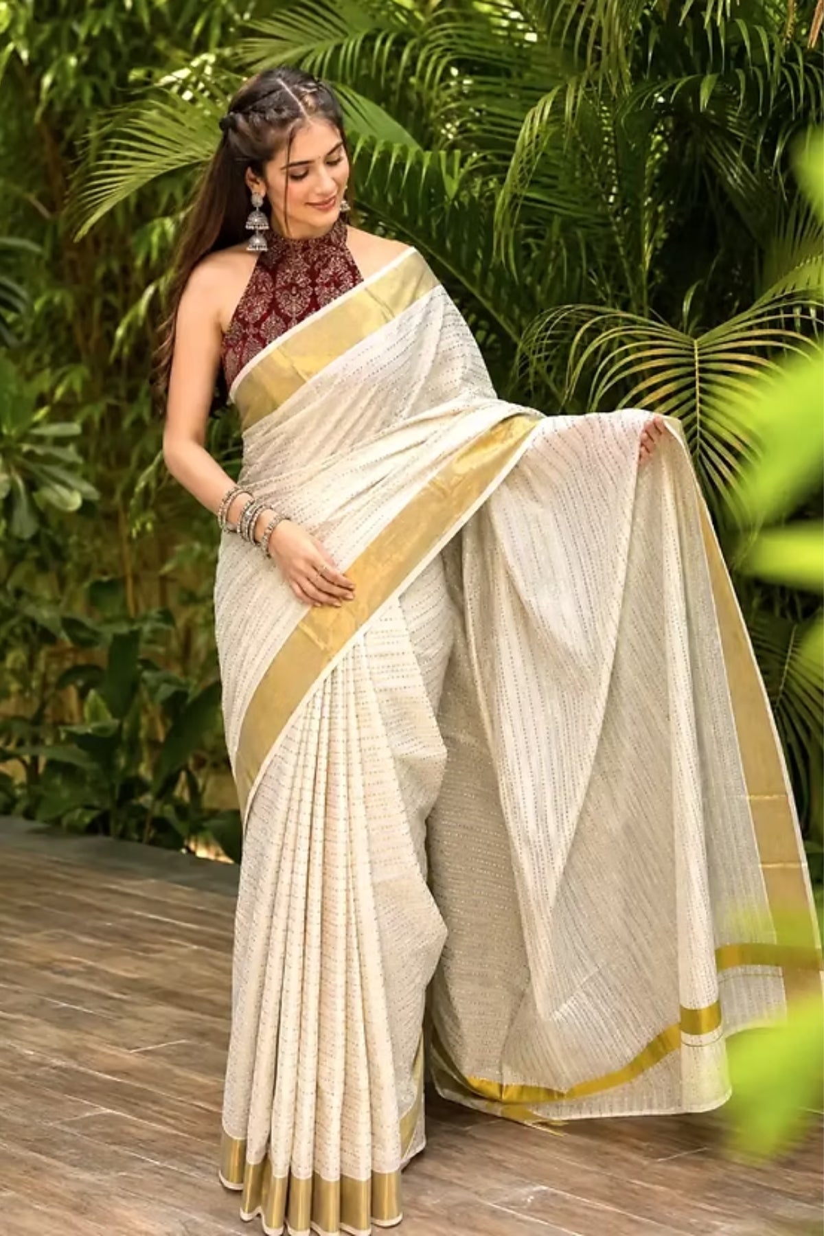 A woman models a cream-colored cotton saree with gold stripes and a matching cream dupatta, paired with a dark red patterned sleeveless blouse, set against a lush green tropical background.