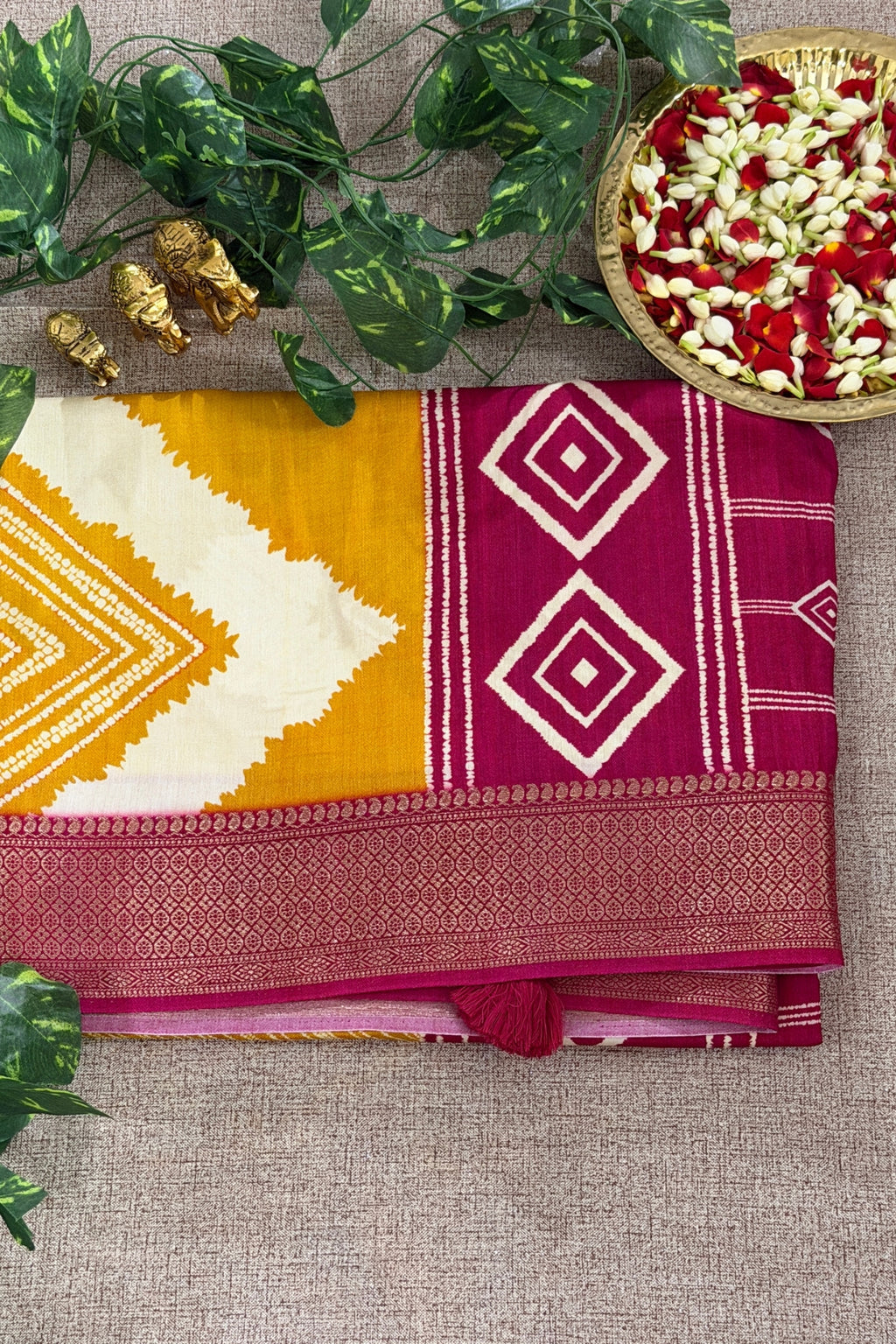 A folded silk saree with a yellow and off-white ikat pattern, a magenta border with cream diamond motifs, and a metallic pink border, styled on a beige fabric background with green leaves and a basket of flowers.
