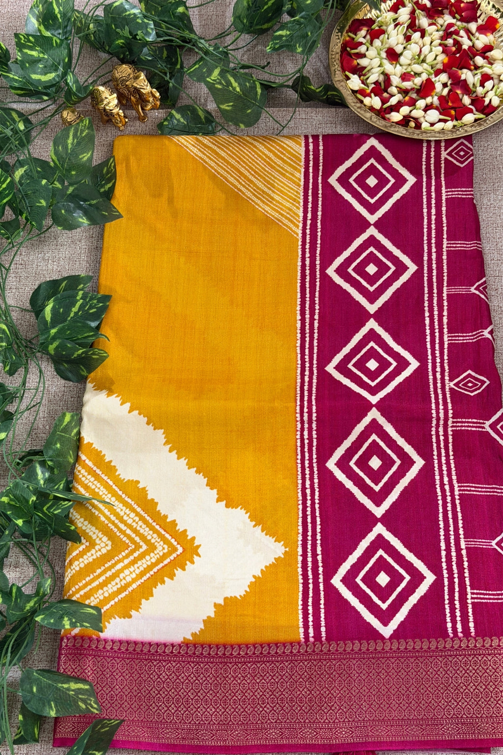 A folded traditional Indian saree displayed flat on a textured beige surface, decorated with green leaves and a bowl of flower petals. The saree features a mustard yellow body, a white chevron pattern, and a magenta border with white diamond motifs.