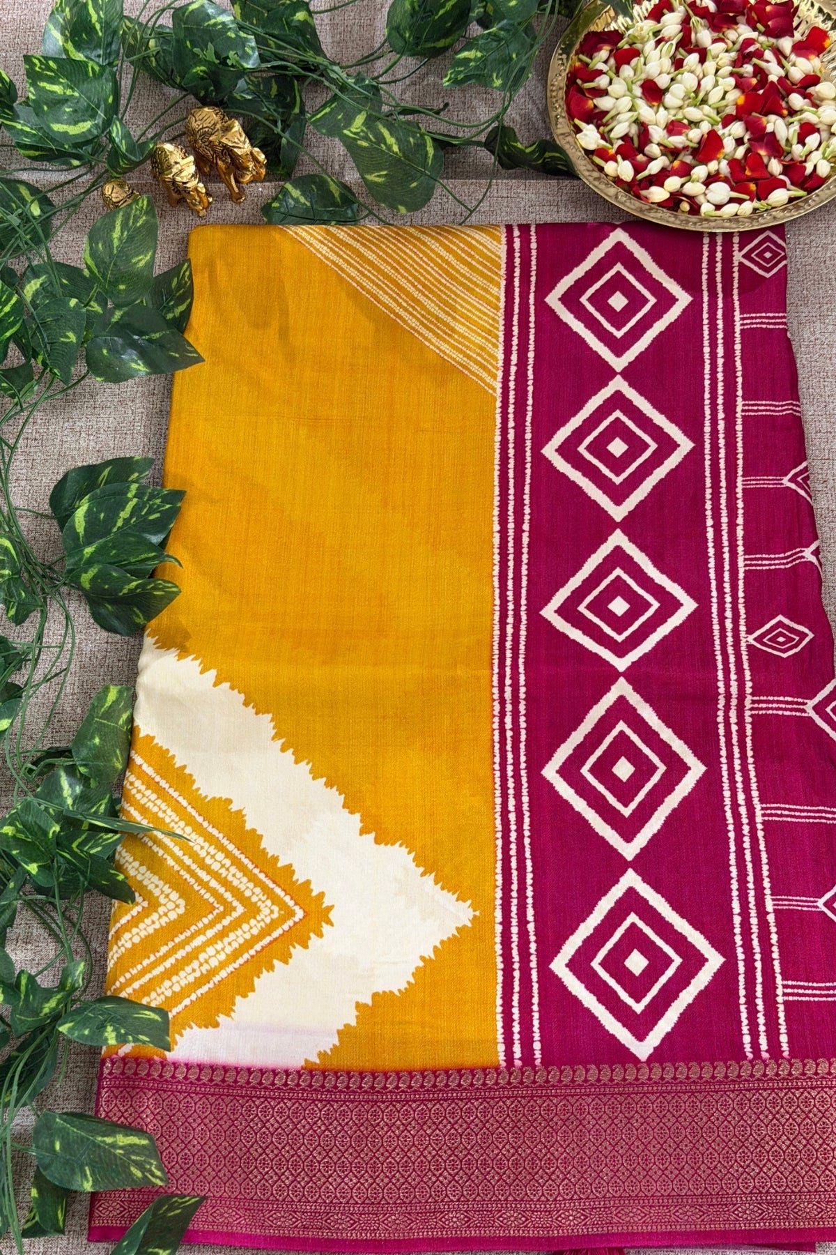 A folded traditional Indian saree displayed flat on a textured beige surface, decorated with green leaves and a bowl of flower petals. The saree features a mustard yellow body, a white chevron pattern, and a magenta border with white diamond motifs.