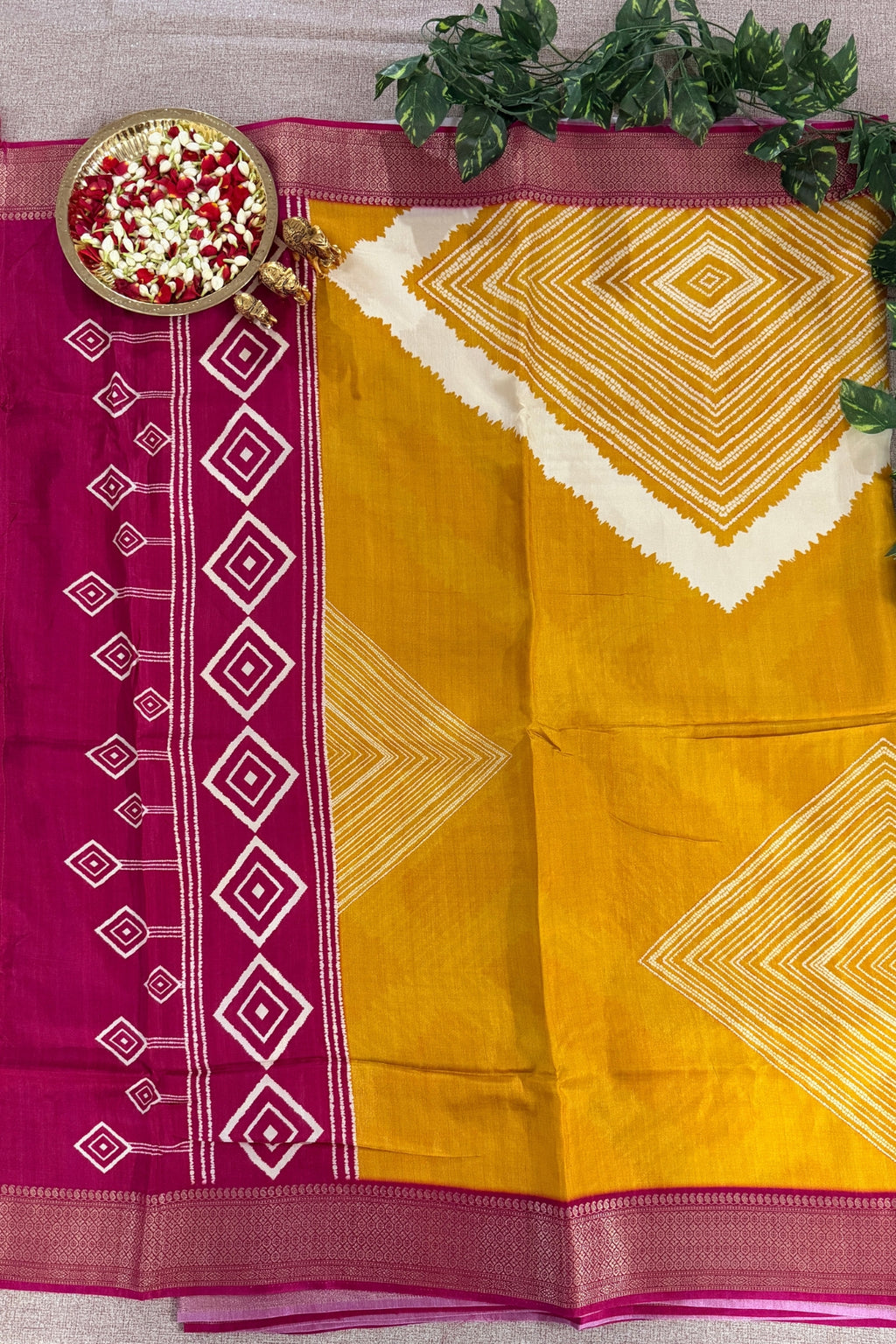 A yellow and magenta saree with white geometric diamond patterns, styled flat-lay with a decorative bowl of flowers and green leaves on a neutral fabric background.