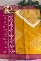 A yellow and magenta saree with white geometric diamond patterns, styled flat-lay with a decorative bowl of flowers and green leaves on a neutral fabric background.