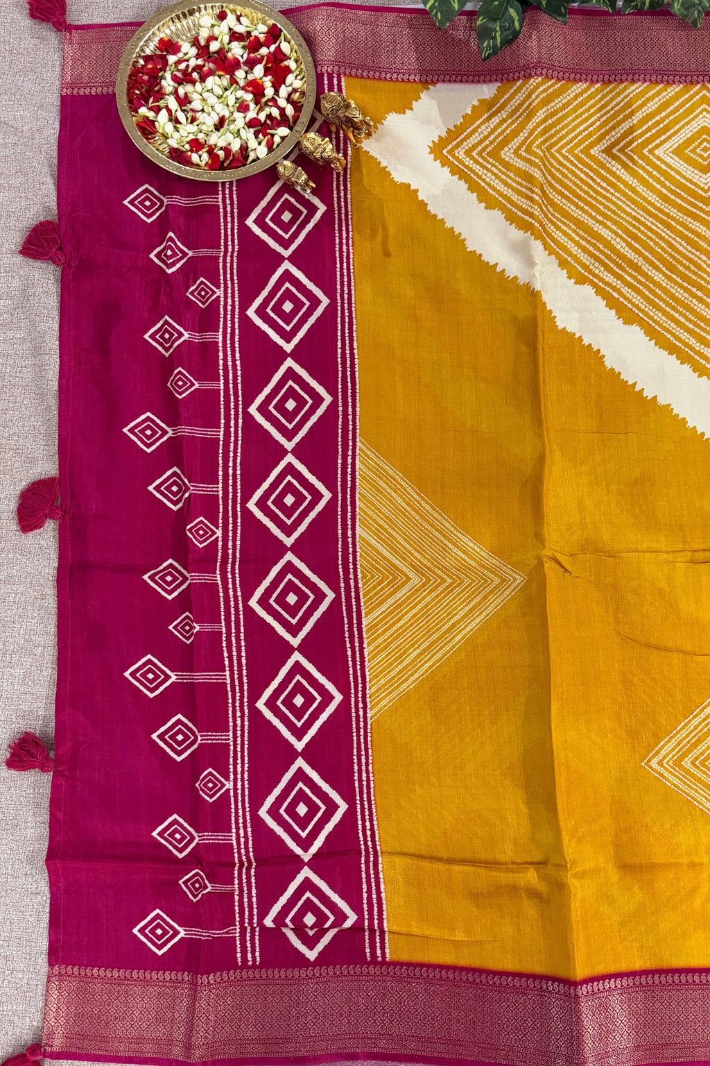 A yellow and magenta Patola-style saree with white geometric patterns, displayed flat with a decorative plate of flowers on a neutral fabric background.