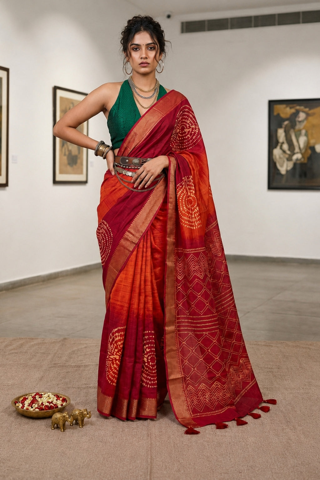 A woman models a red and orange saree with intricate gold and red patterns, paired with a green sleeveless blouse, standing in a gallery with framed art on the walls.