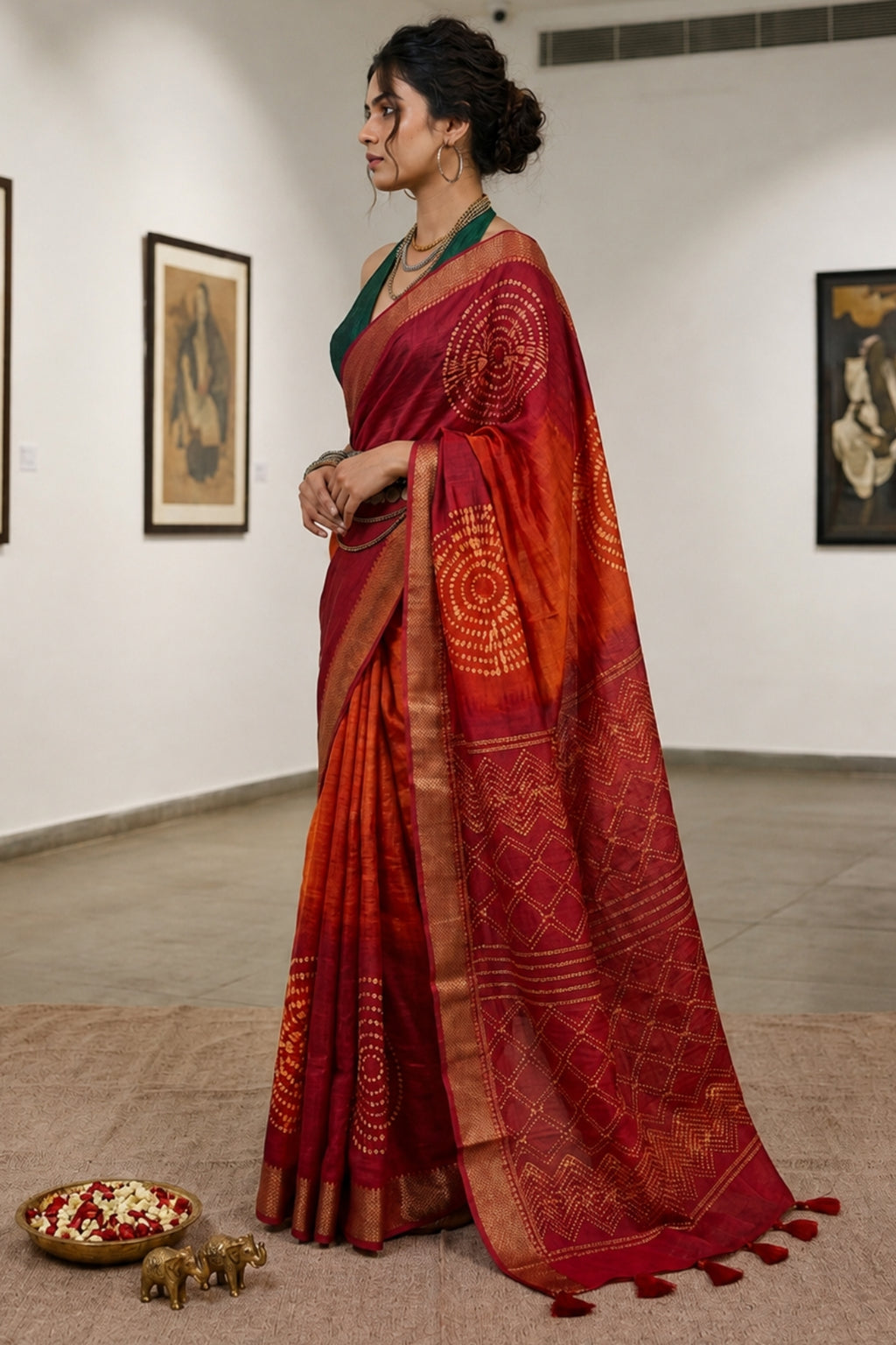 A woman wearing a red and orange saree with gold zari patterns stands in a gallery setting with framed art on the walls. The saree features a deep red border with gold detailing and tassels, and she is wearing a green blouse.