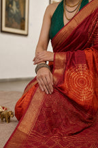 A close-up of a woman wearing a rich red and orange saree with intricate gold zari patterns and a green blouse, seated indoors.