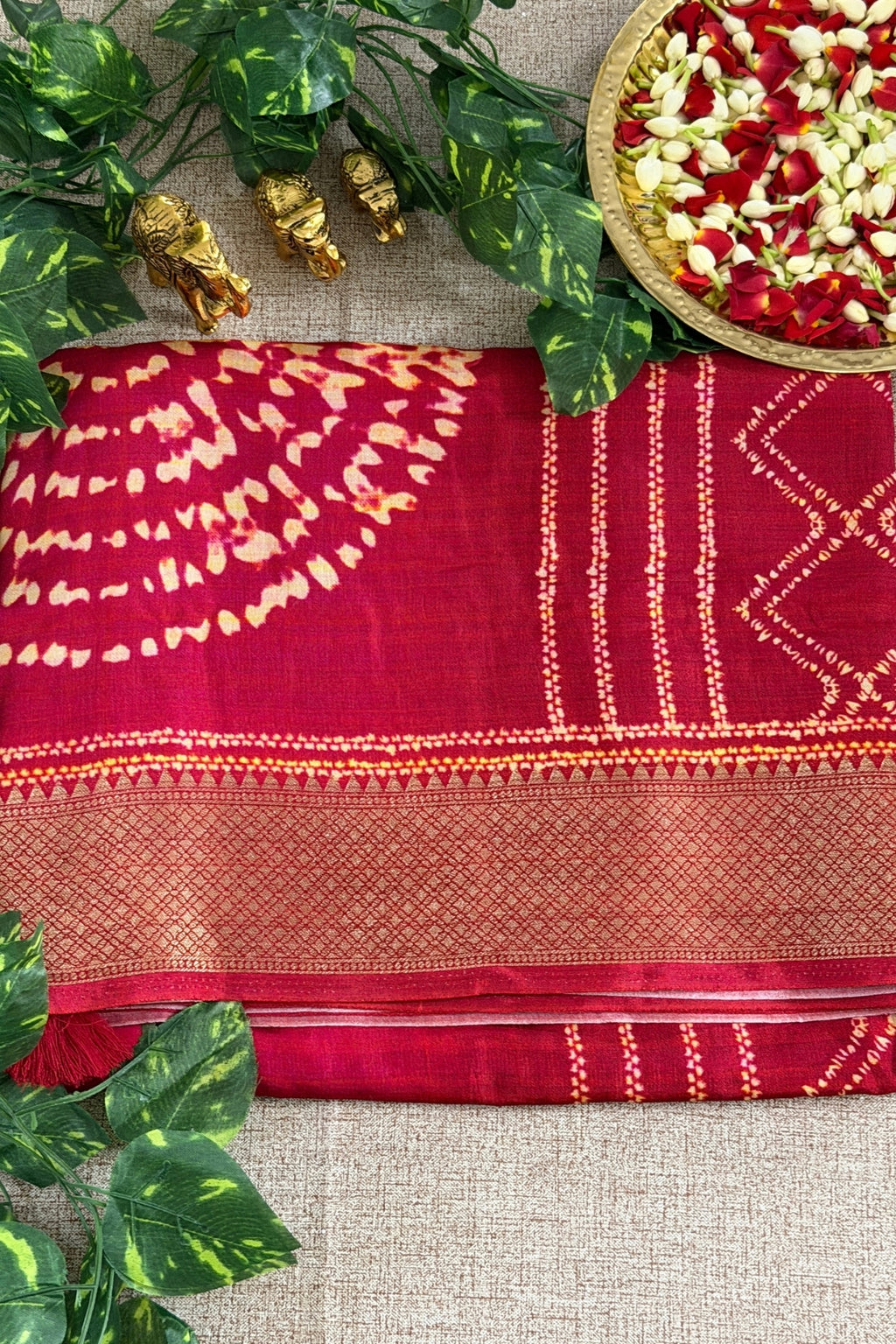 A folded deep red saree with gold zari embroidery and tie-dye patterns, styled on a beige textured fabric background with decorative green leaves, small gold figurines, and a plate of flower petals.