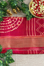 A folded deep red saree with gold zari embroidery and tie-dye patterns, styled on a beige textured fabric background with decorative green leaves, small gold figurines, and a plate of flower petals.