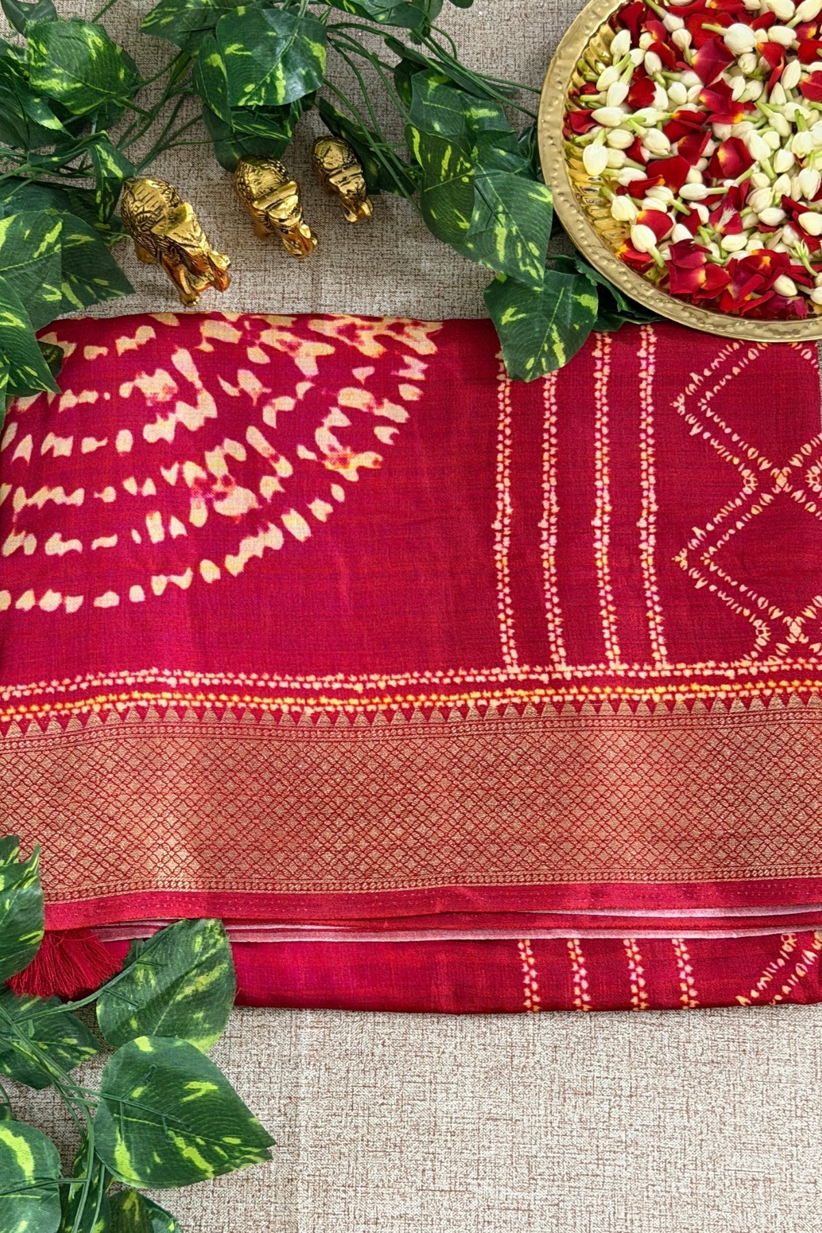 A folded deep red saree with gold zari embroidery and tie-dye patterns, styled on a beige textured fabric background with decorative green leaves, small gold figurines, and a plate of flower petals.