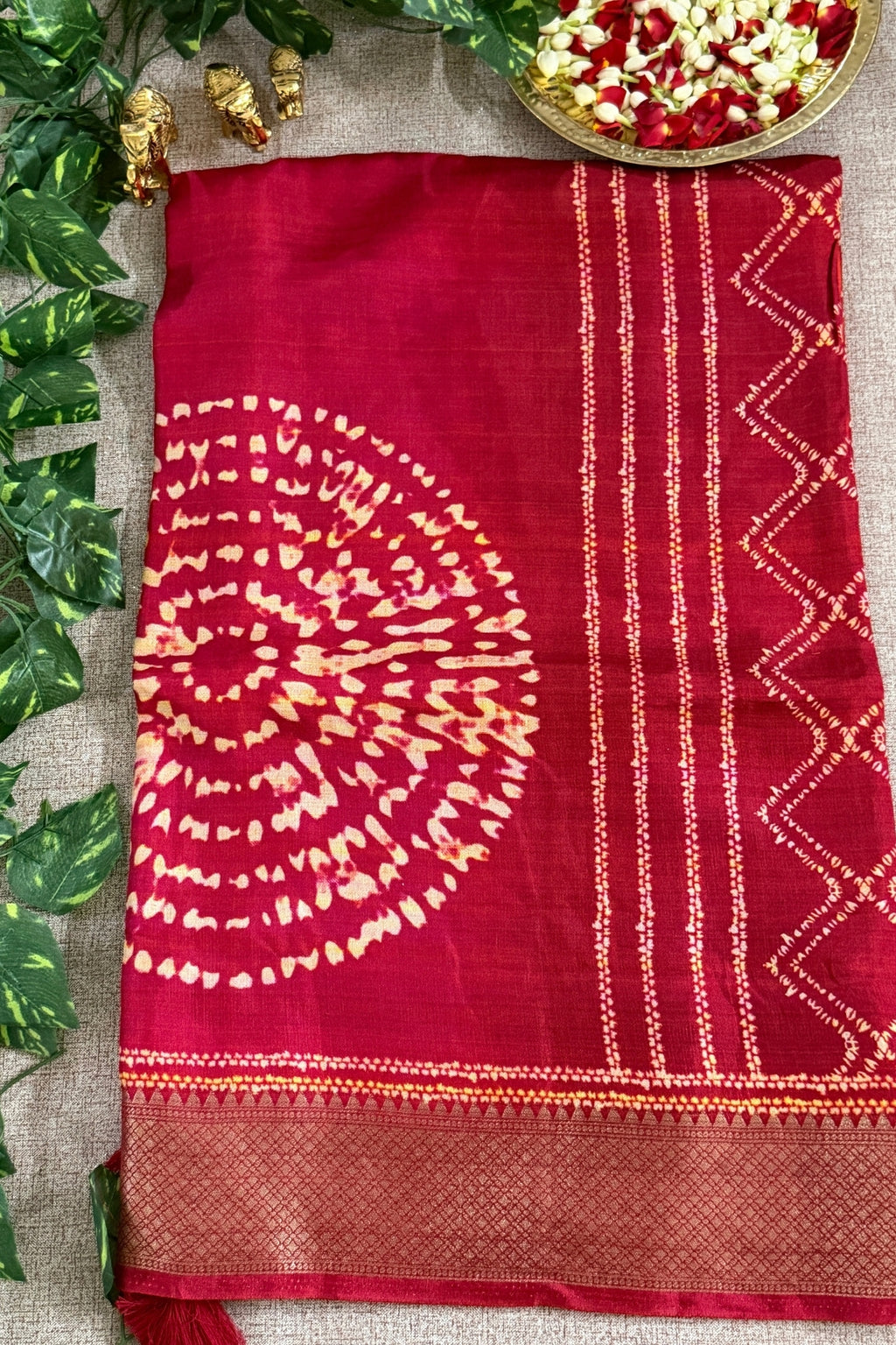 A deep red silk saree with a gold zari printed circular motif and geometric borders, laid flat on a beige burlap surface with decorative green leaves and a gold bowl of flowers.