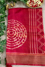 A deep red silk saree with a gold zari printed circular motif and geometric borders, laid flat on a beige burlap surface with decorative green leaves and a gold bowl of flowers.