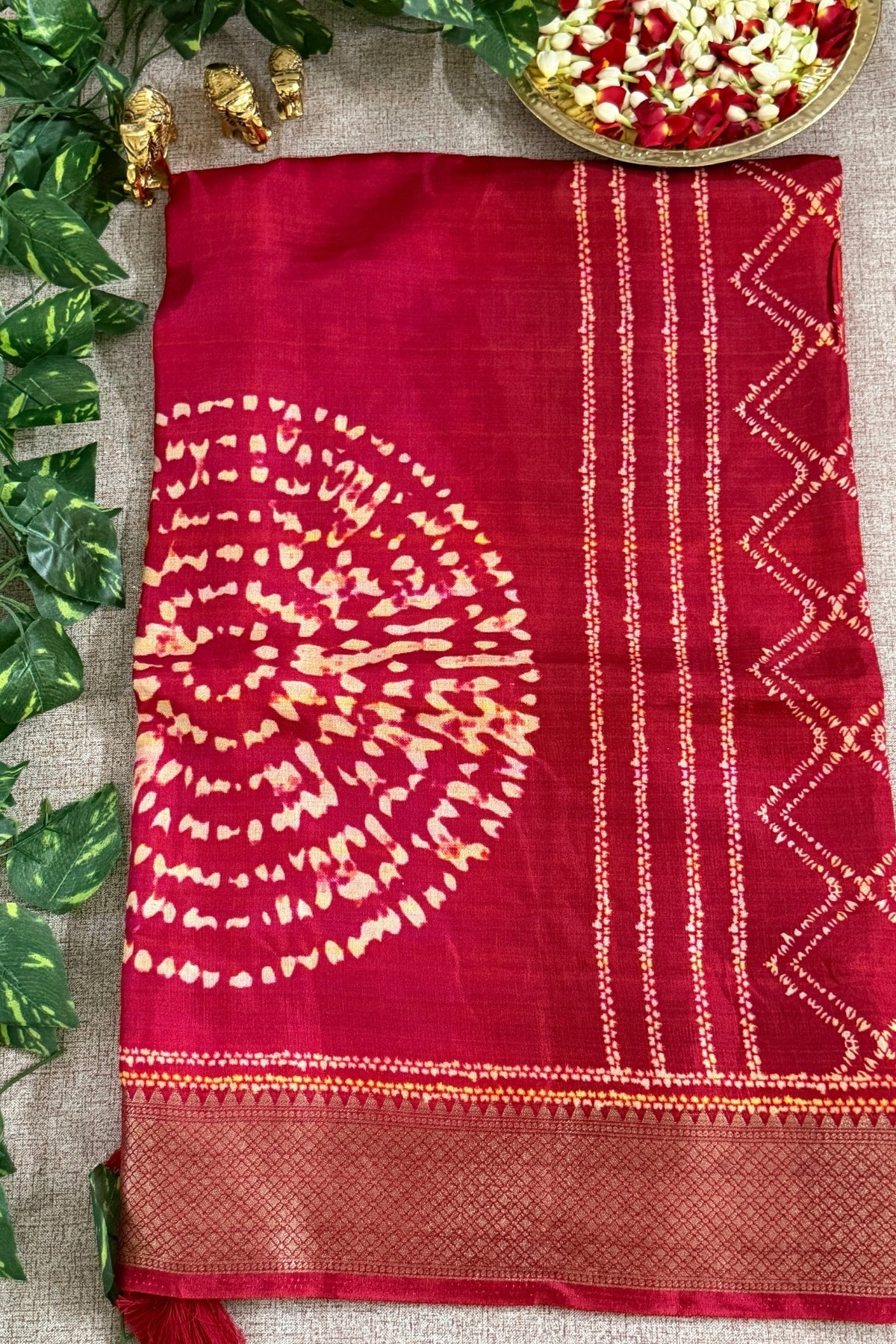 A deep red silk saree with a gold zari printed circular motif and geometric borders, laid flat on a beige burlap surface with decorative green leaves and a gold bowl of flowers.