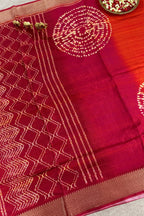 A close-up of a deep red or maroon silk saree with intricate gold zari embroidery, featuring a geometric diamond pattern on the border, vertical stripes, and circular motifs on the body. The fabric is draped elegantly, showing its texture and sheen.