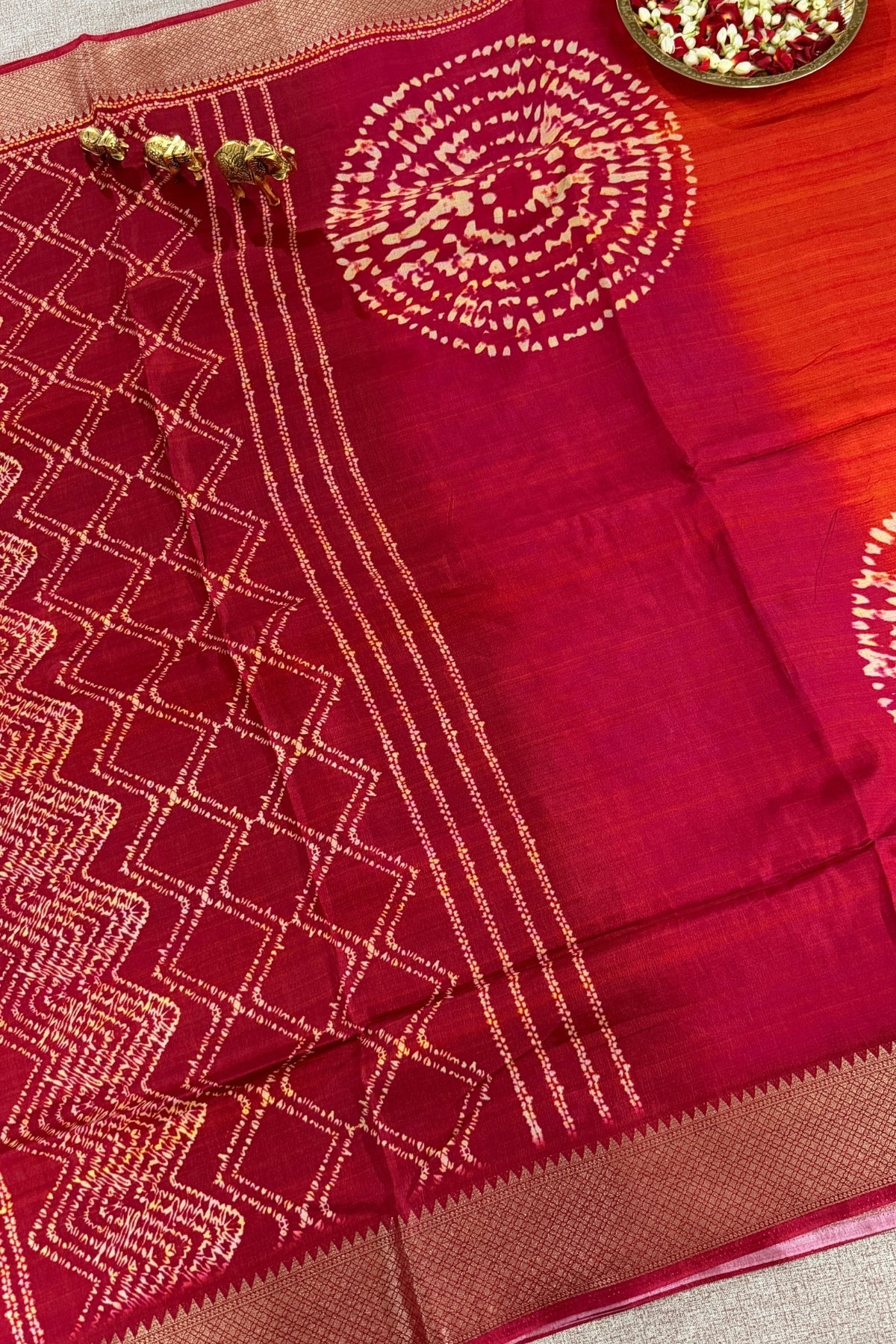 A close-up of a deep red or maroon silk saree with intricate gold zari embroidery, featuring a geometric diamond pattern on the border, vertical stripes, and circular motifs on the body. The fabric is draped elegantly, showing its texture and sheen.